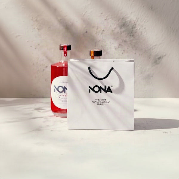 A white NONA paper shopping bag stands in front of a clear bottle filled with pink liquid on a light marble surface against a textured gray wall with soft shadows.