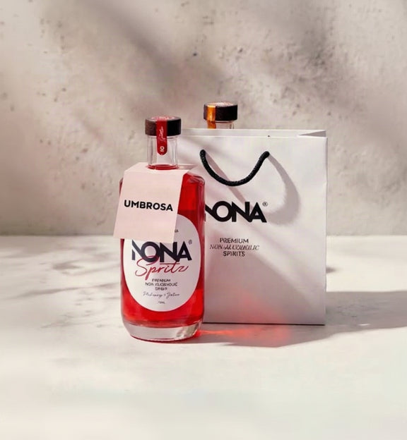 A bottle of Nona Spiritz premium non-alcoholic spirits in a clear glass bottle with red liquid, shown next to a white Nona gift bag on a light marble surface against a textured concrete wall with soft shadows.